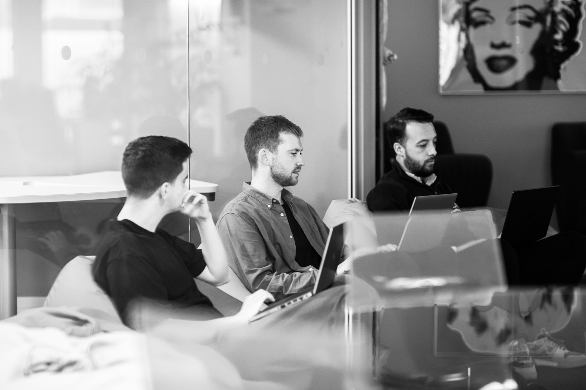 Three men working on laptops in modern office meeting room with glass walls and artwork on wall