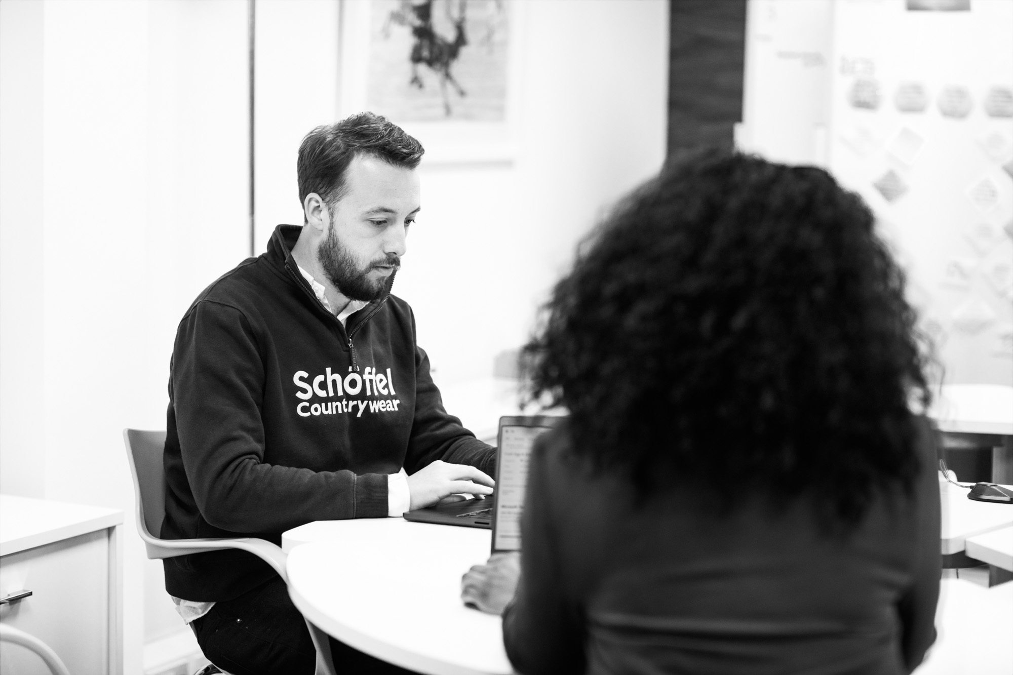 Man in Schöffel sweatshirt working on laptop while meeting with colleague in modern office space