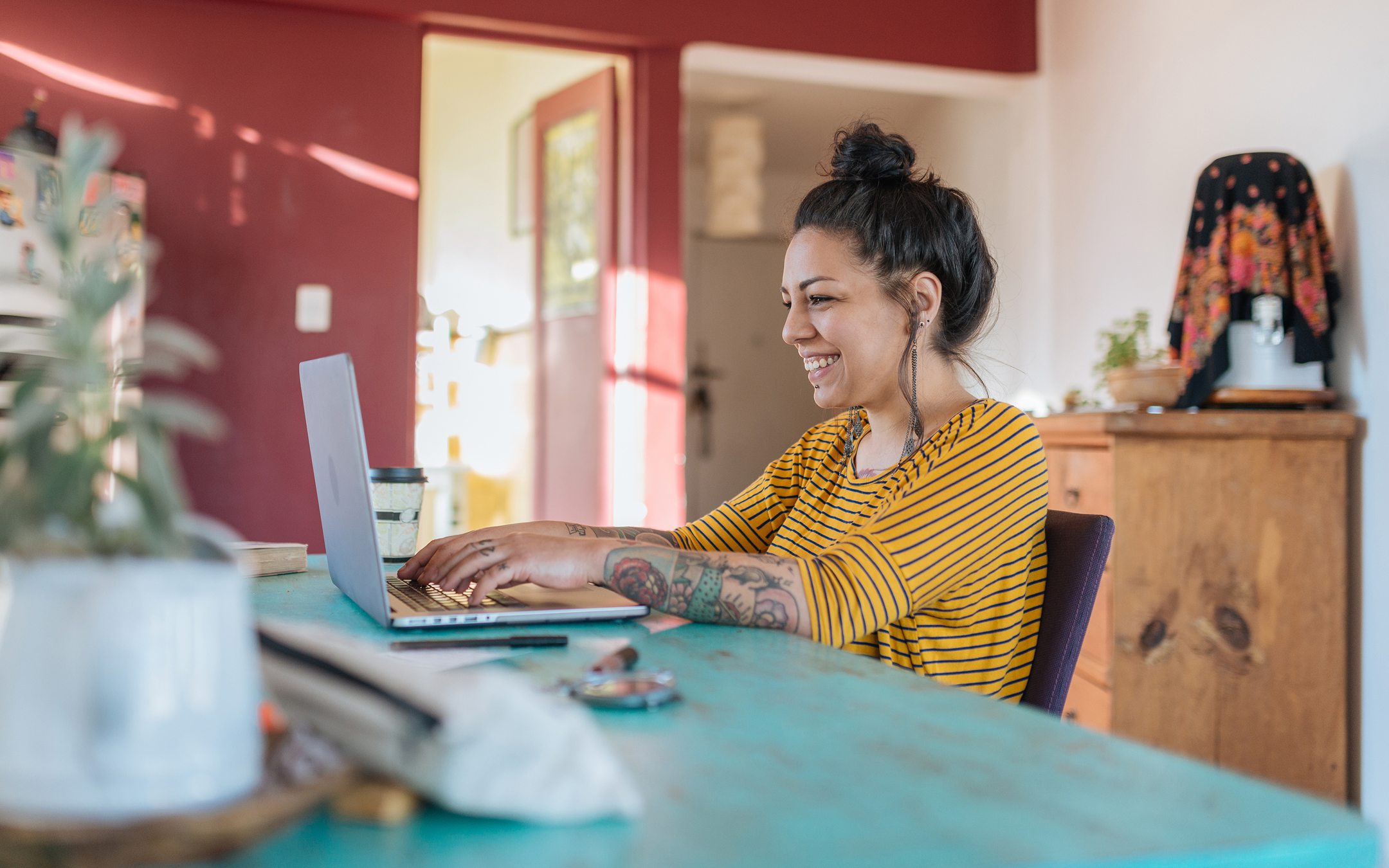 Woman with tattoos working on laptop at turquoise table in colorful home office wearing yellow striped shirt