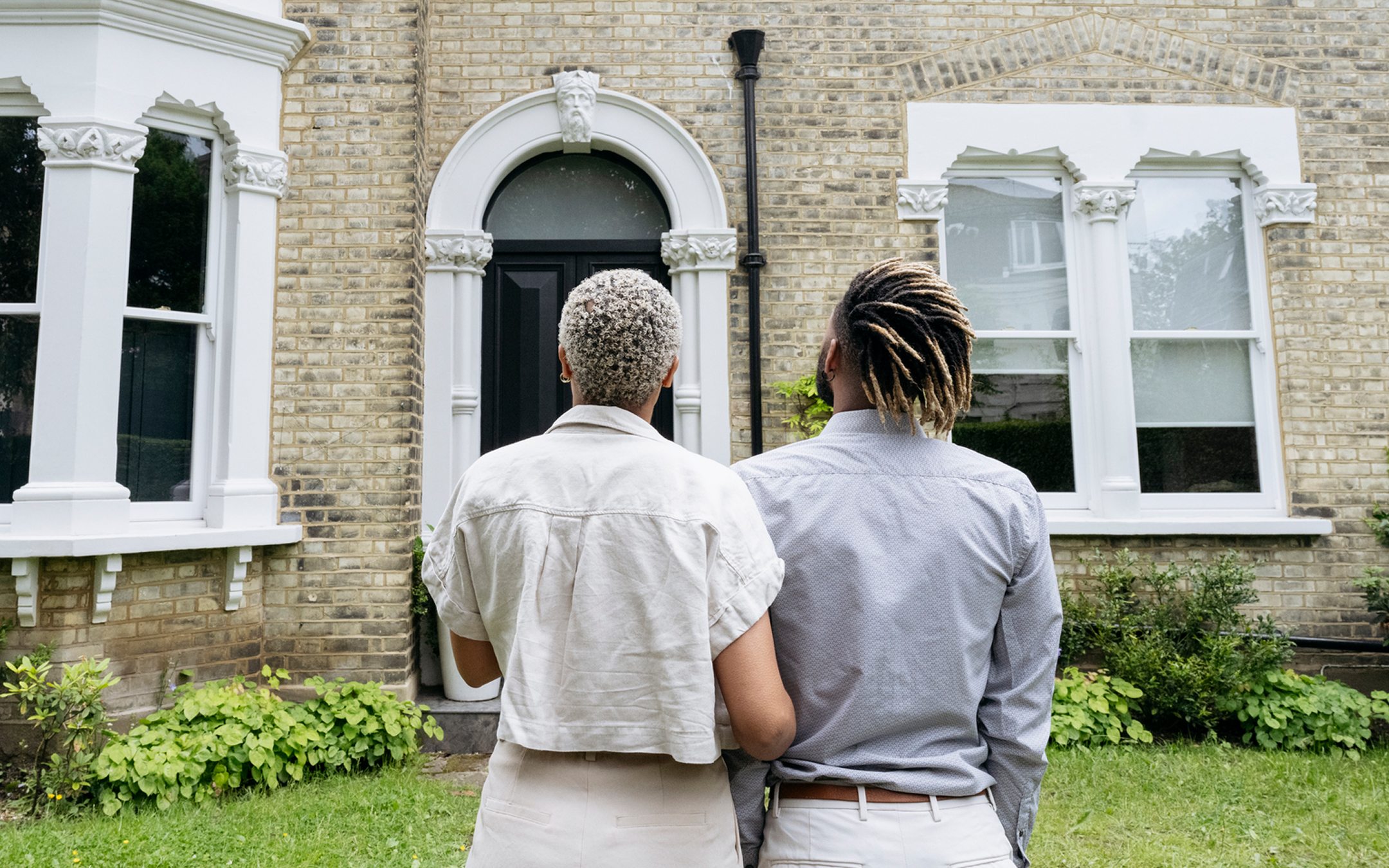 Couple standing together looking at Victorian brick house with white architectural details and front entrance