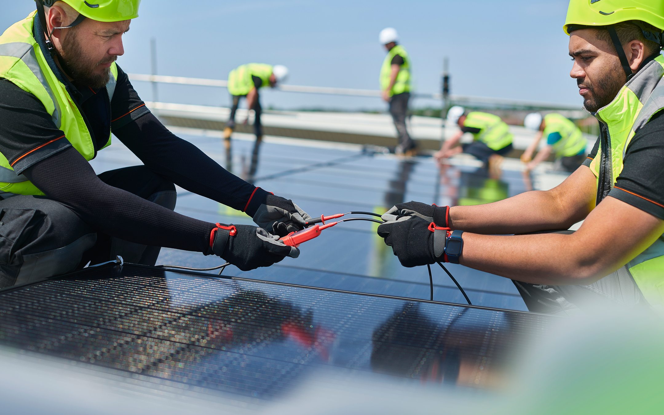 Two workers in high-visibility safety gear installing solar panels on a rooftop with blue sky in background