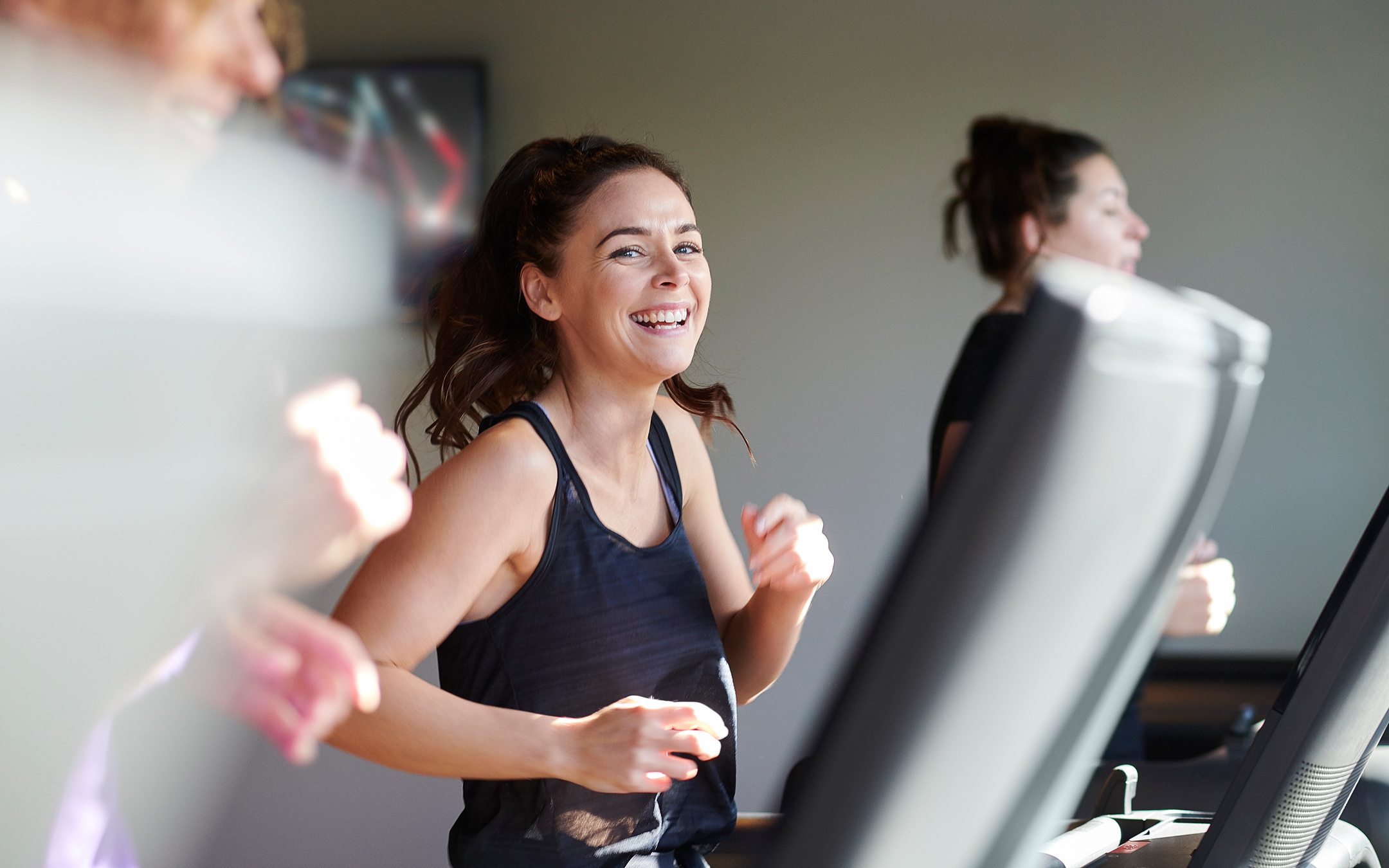 Woman smiling while running on treadmill in gym wearing navy tank top with other people exercising in background