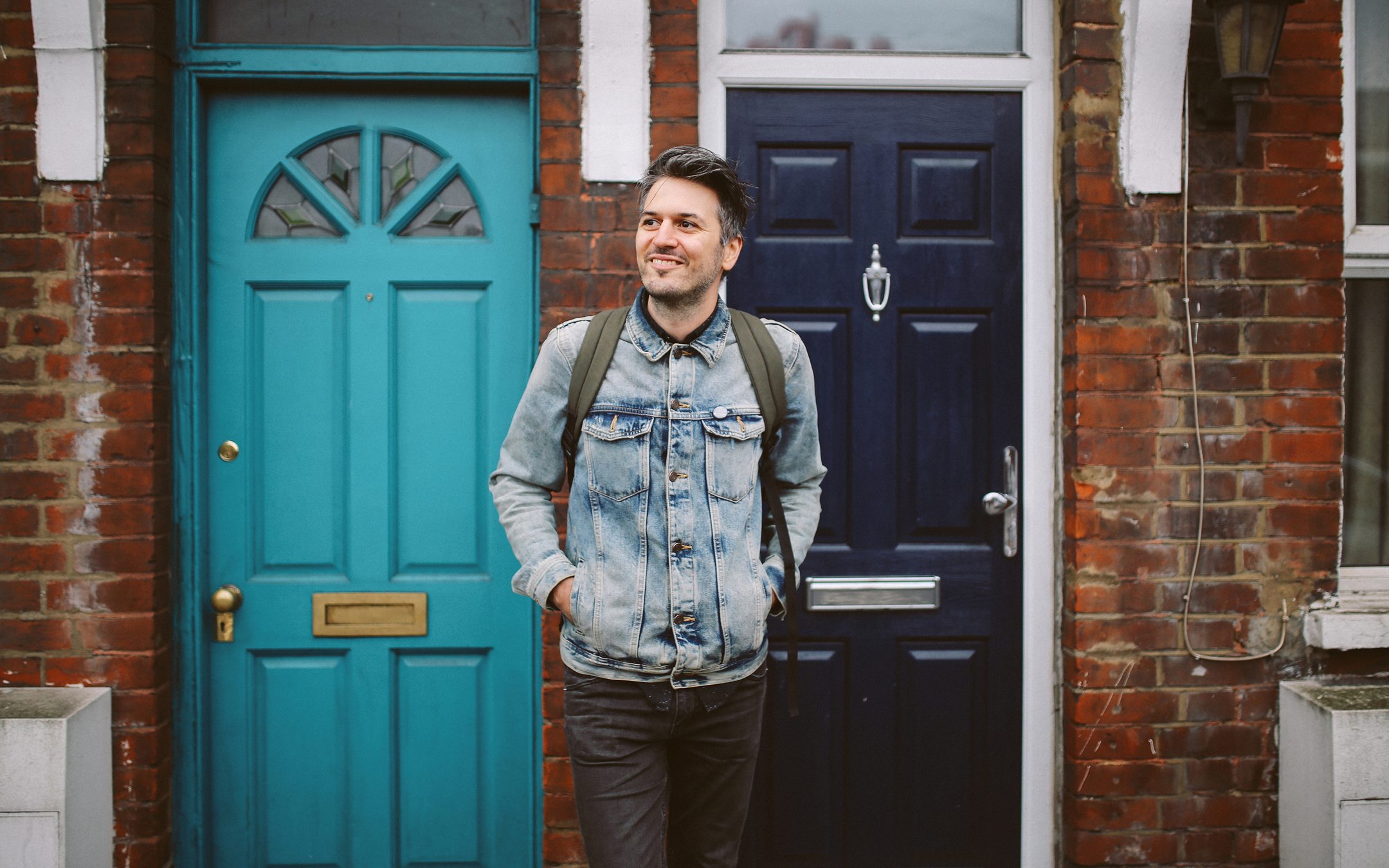 Young man in denim jacket and backpack standing outdoors in front of colorful brick townhouse doors