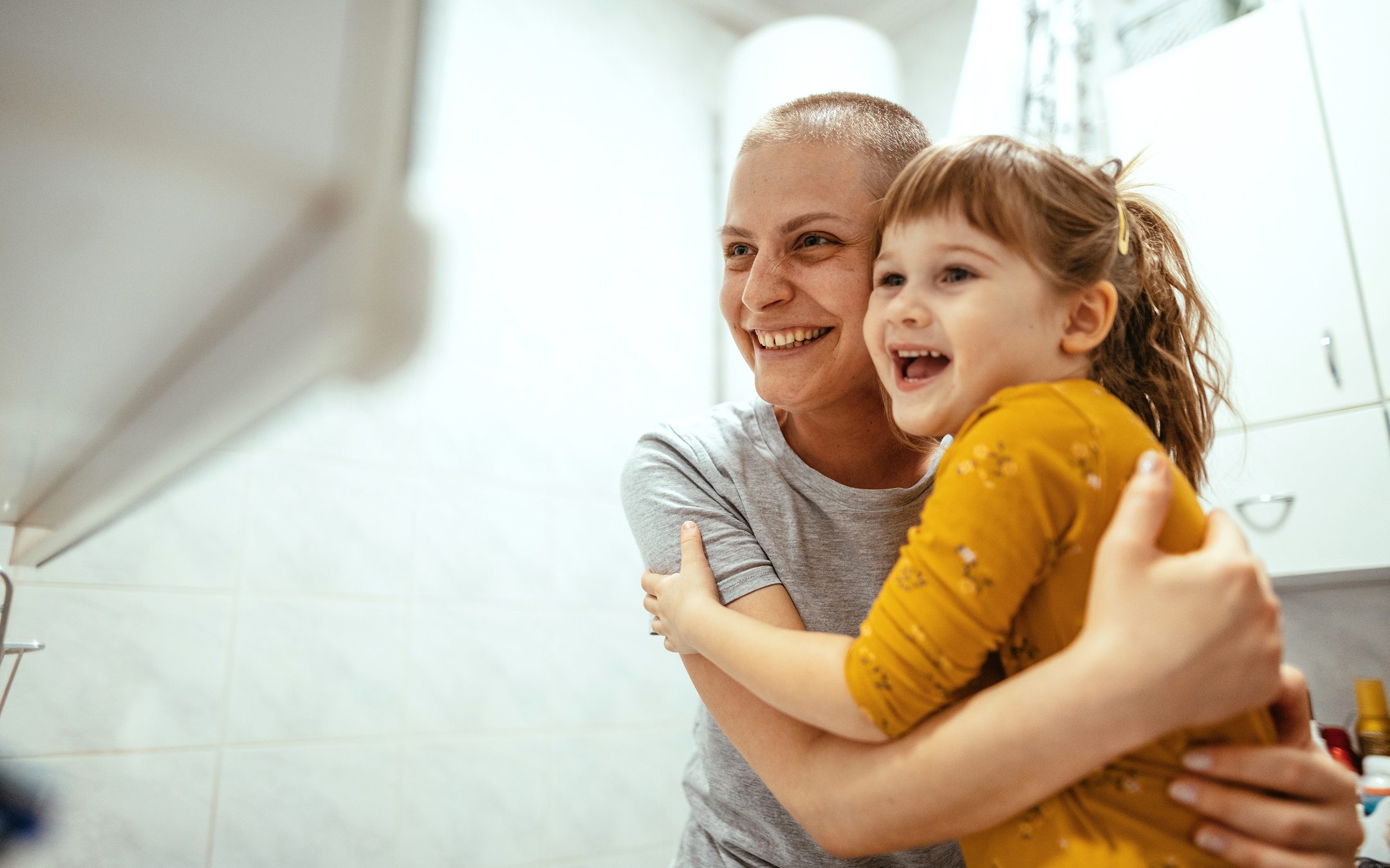 Smiling woman with short hair embracing young girl in yellow shirt in bright kitchen setting