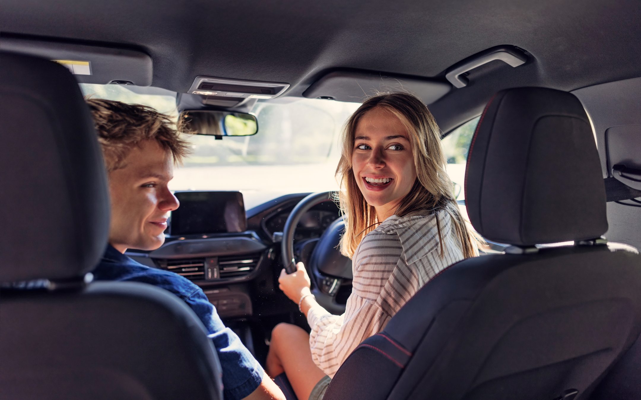 Two young people smiling and talking inside a car, woman in passenger seat wearing striped sweater
