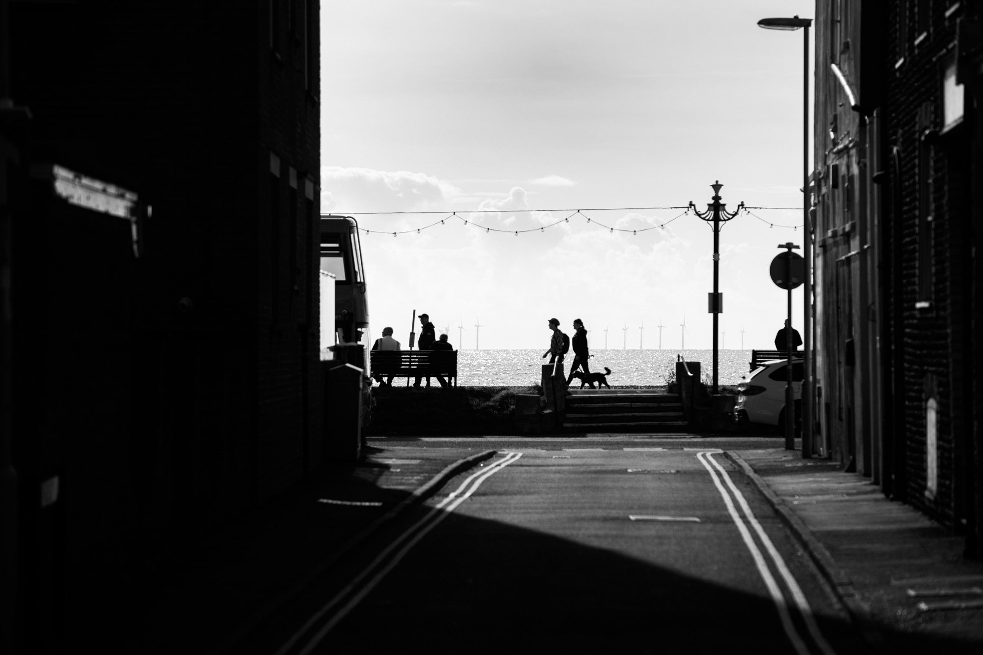Black and white street scene showing narrow road leading to waterfront with silhouetted people and ornate lamppost