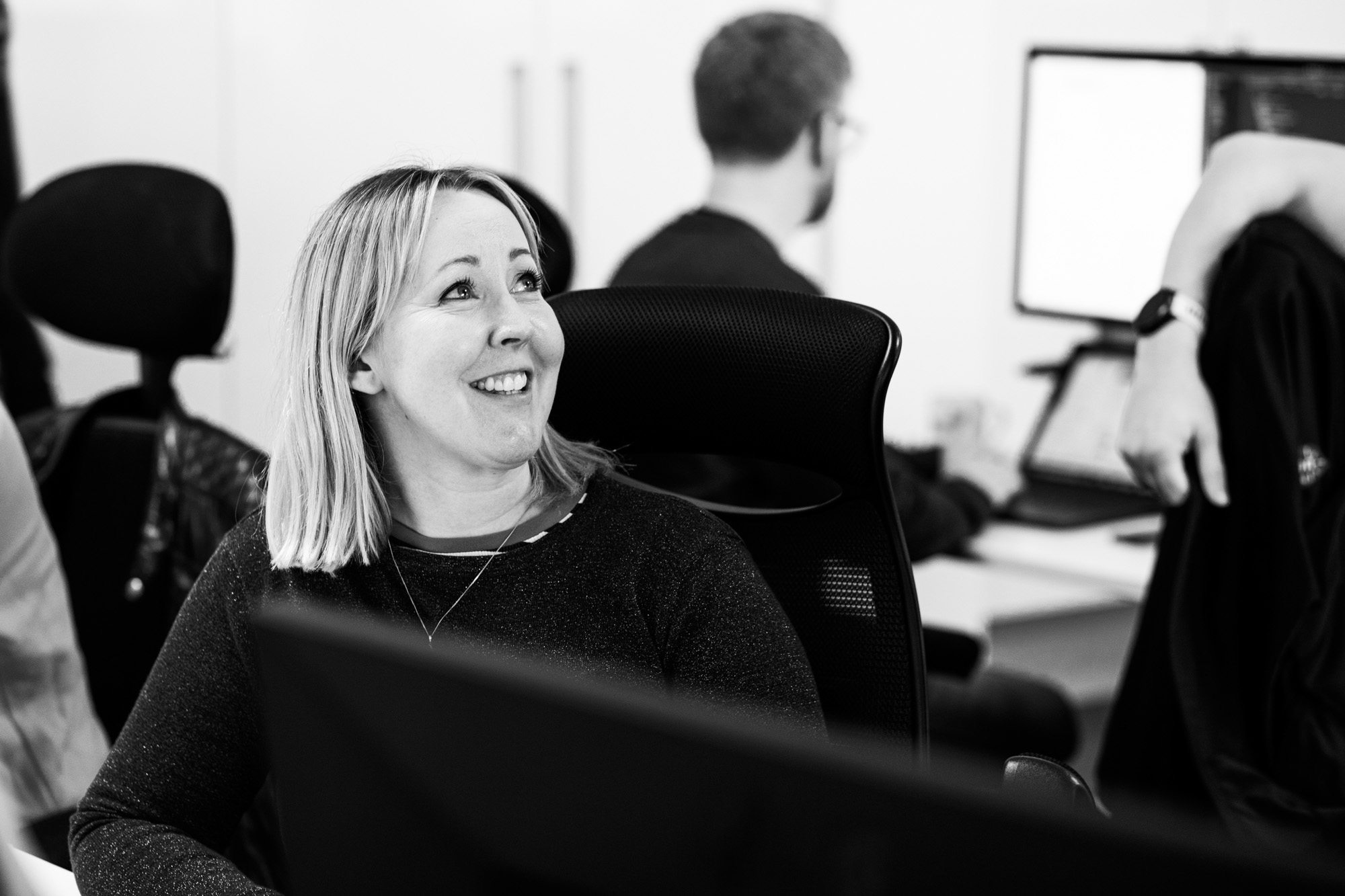 Woman smiling while sitting at desk in modern office with colleagues working in background, black and white photo