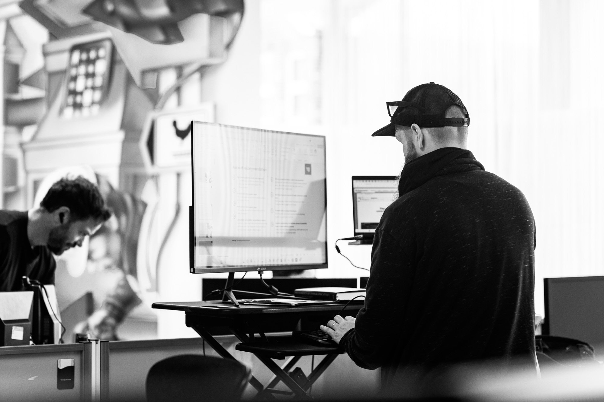 Two people working at standing desks with computers in a modern office environment, black and white photo