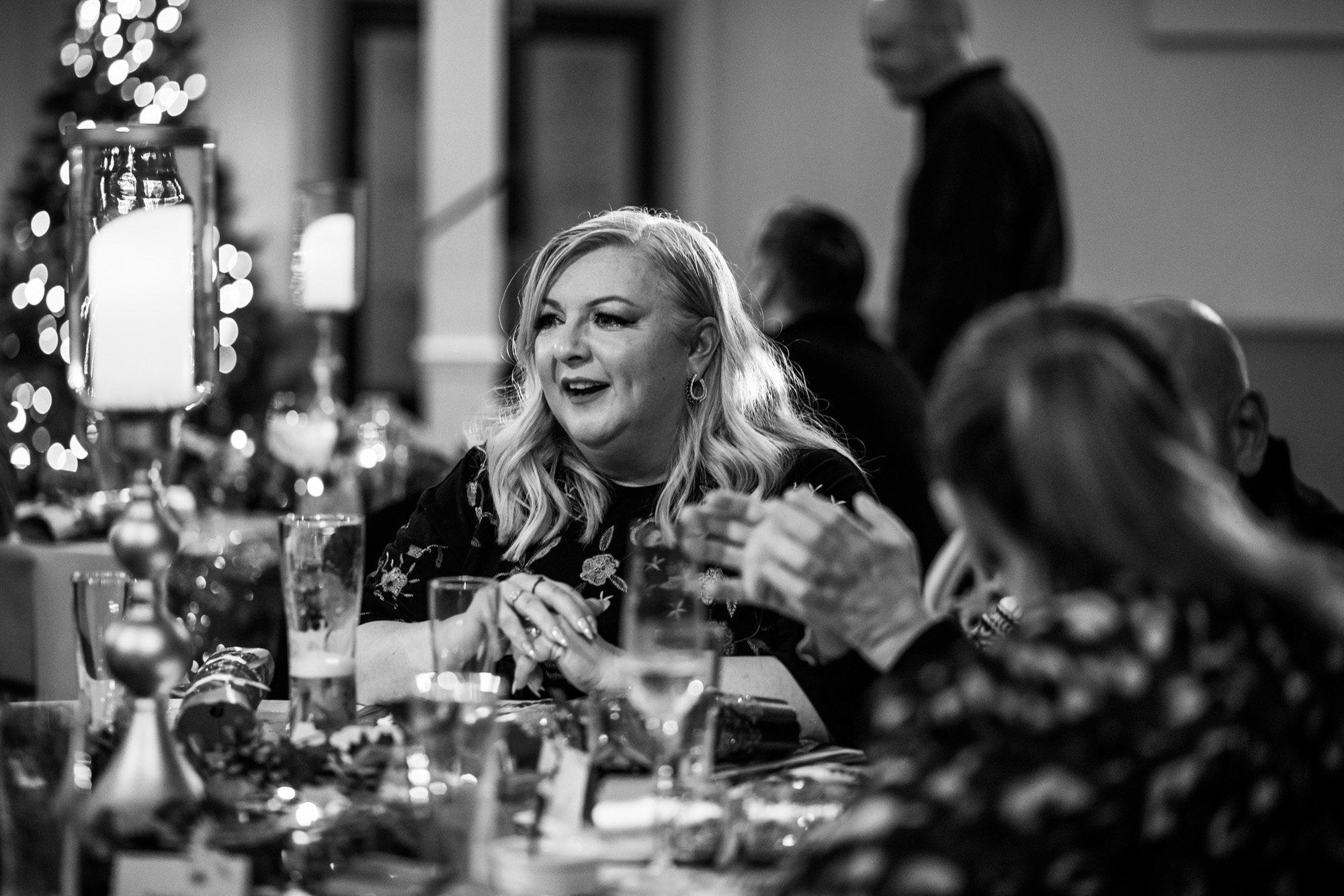Black and white photo of blonde woman smiling at dinner table with wine glasses and festive lighting in background