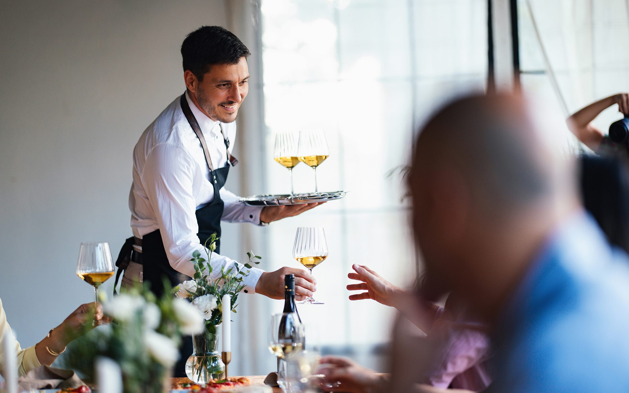 Waiter in white shirt and apron serving white wine to restaurant customers at dining table