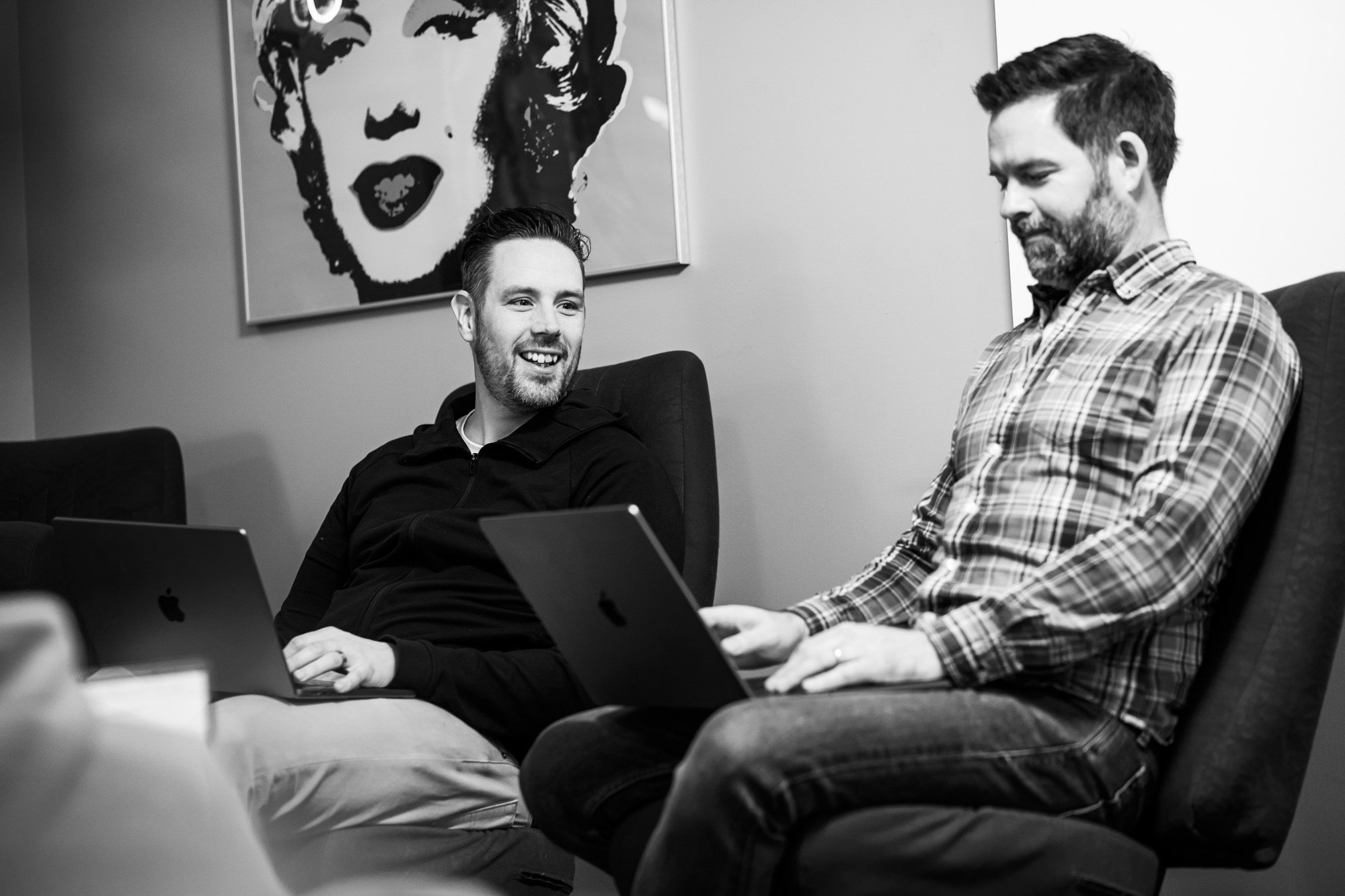 Two men sitting on a couch with laptops in a modern office setting with wall art