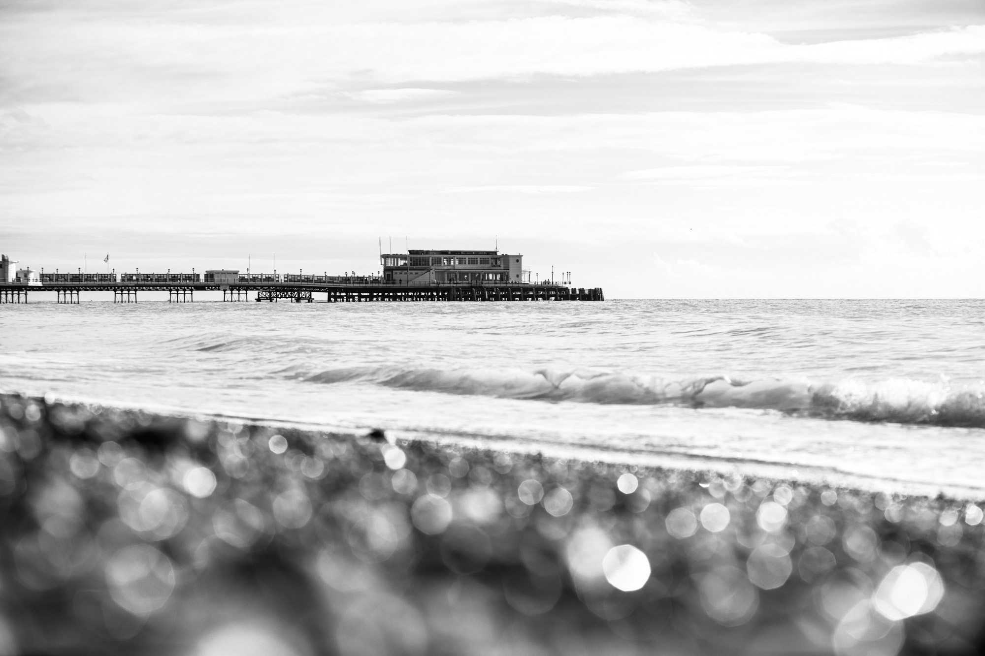 Black and white coastal scene with wooden pier extending into ocean, waves breaking on shore with bokeh light effects