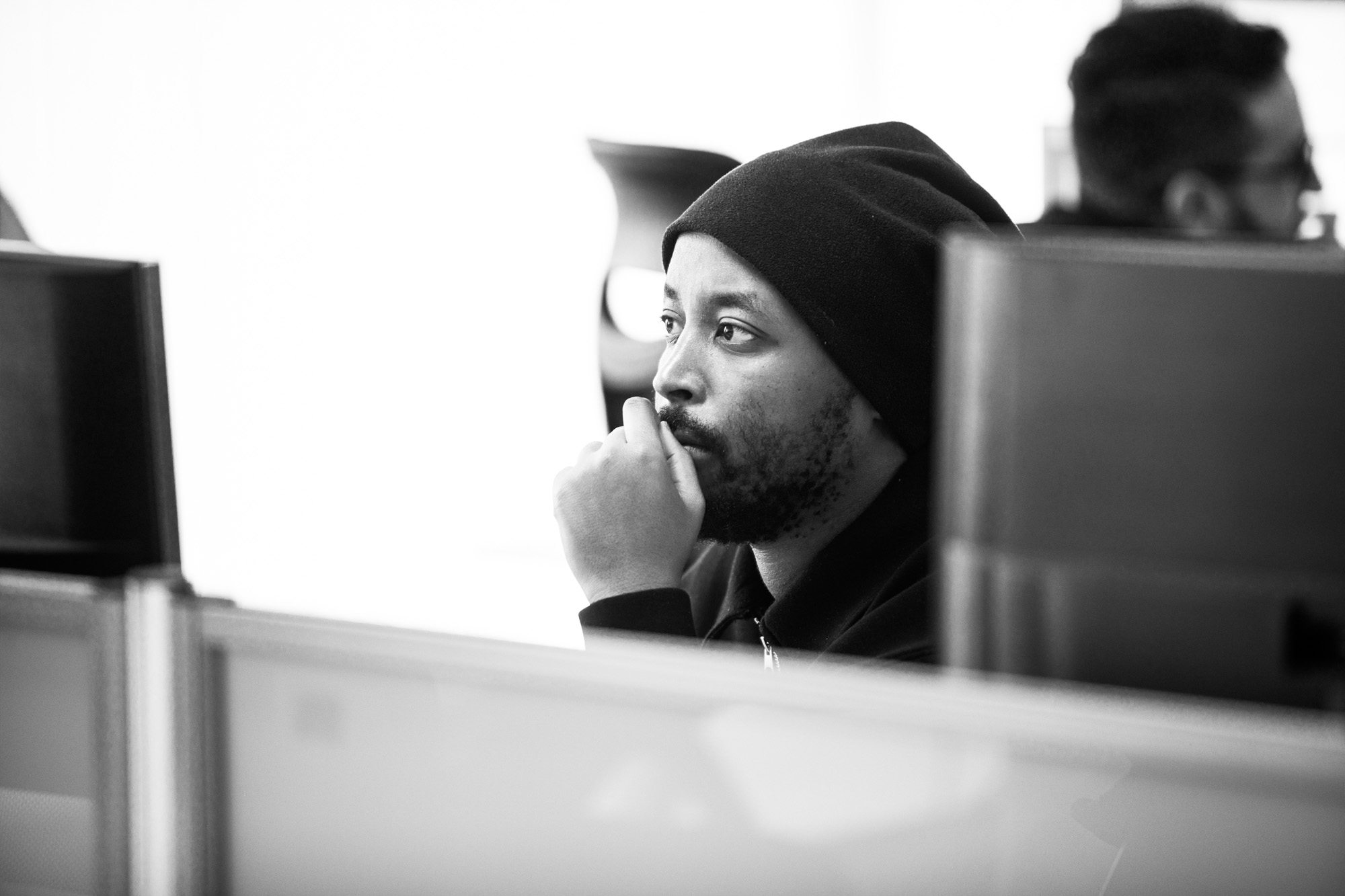 Person in beanie and hoodie sitting at desk in office environment, hand thoughtfully positioned near face