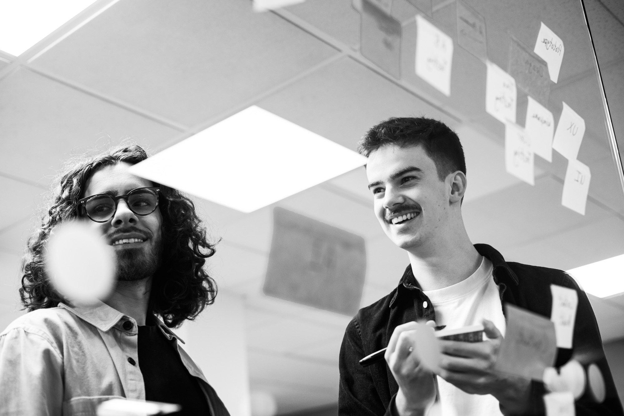 Two young men smiling and looking upward while holding smartphones in an office environment