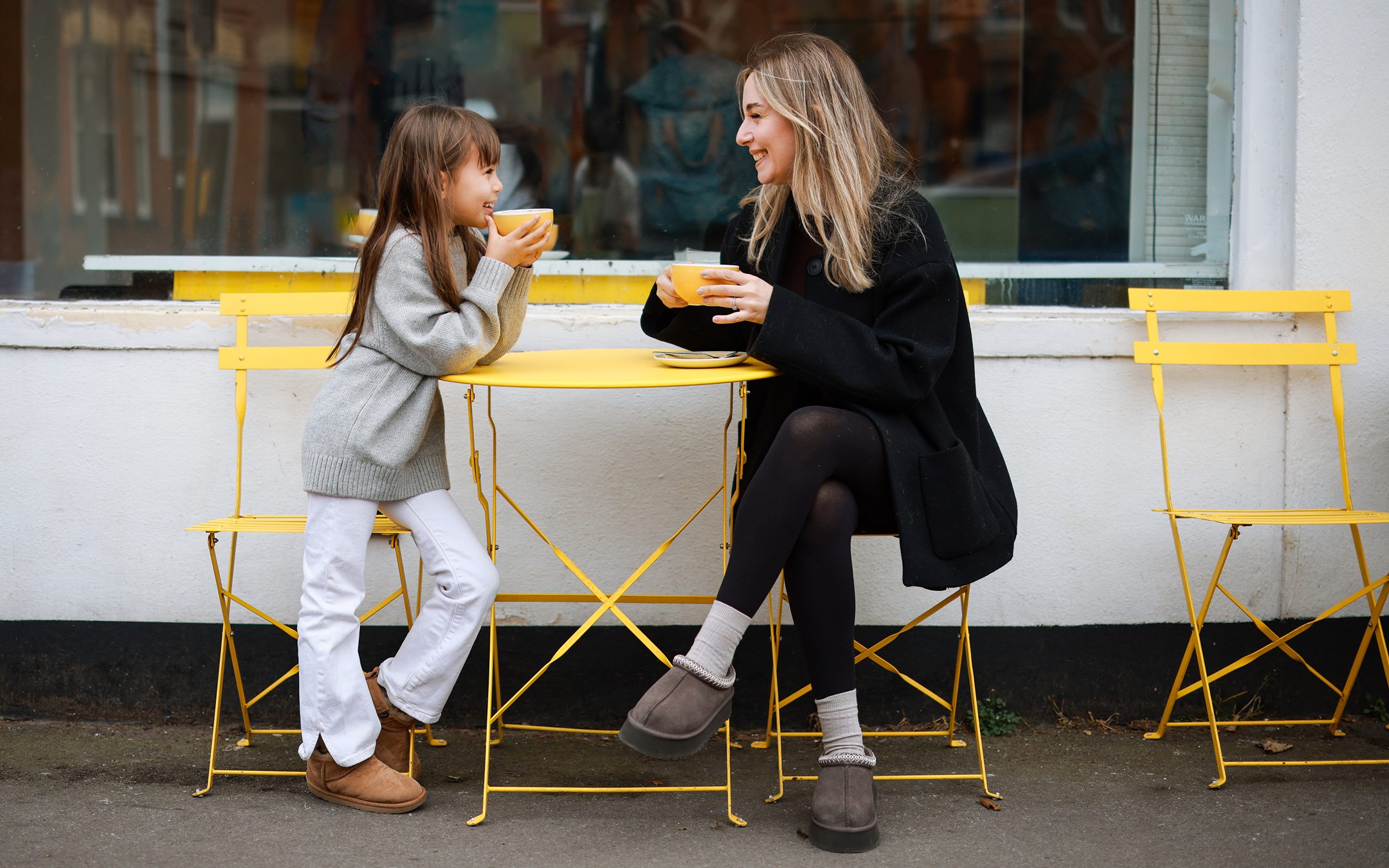 Mother and daughter sharing drinks at yellow outdoor cafe table, smiling and enjoying quality time together