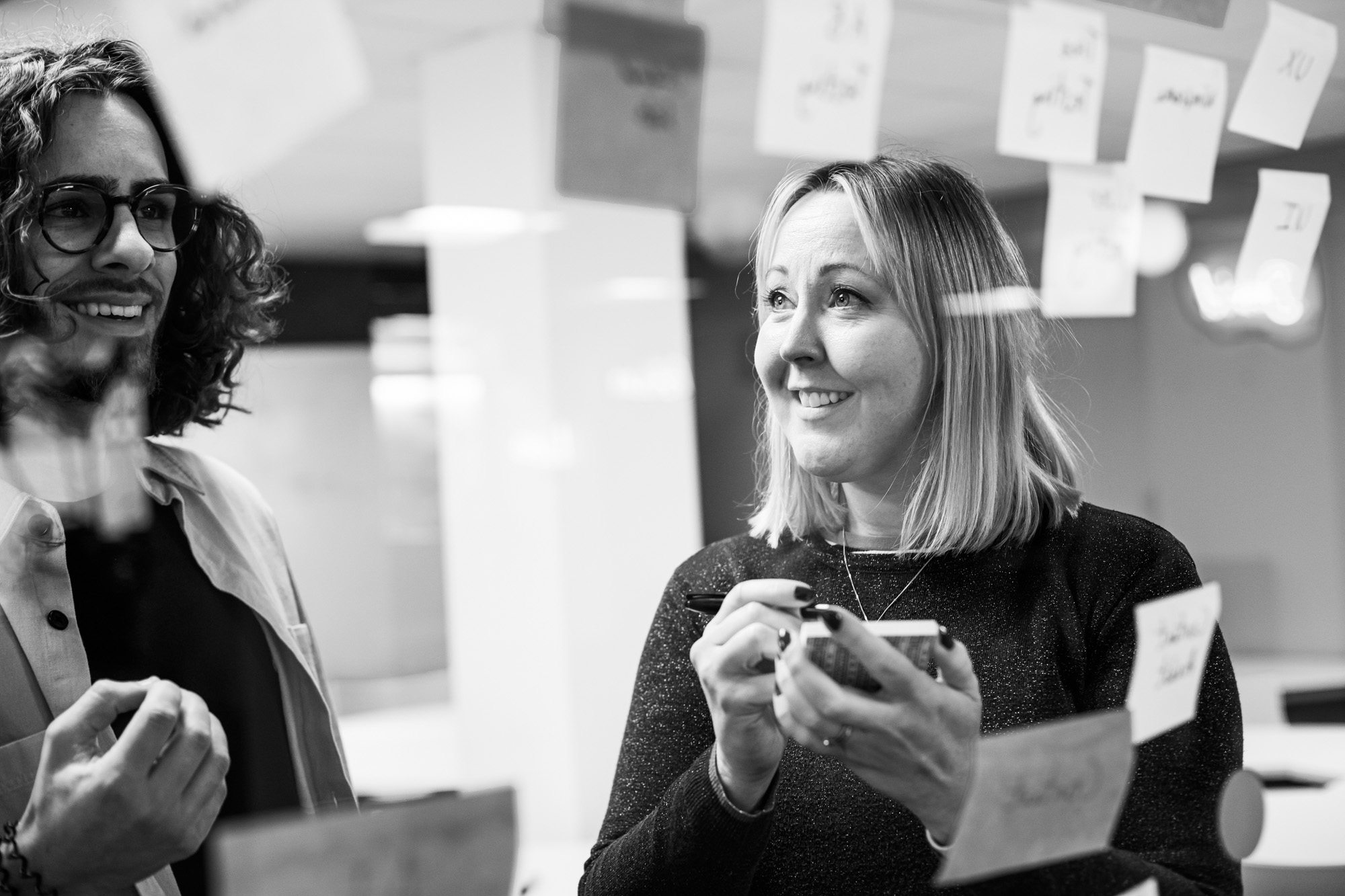 Two smiling professionals collaborating in modern office with sticky notes on glass wall behind them