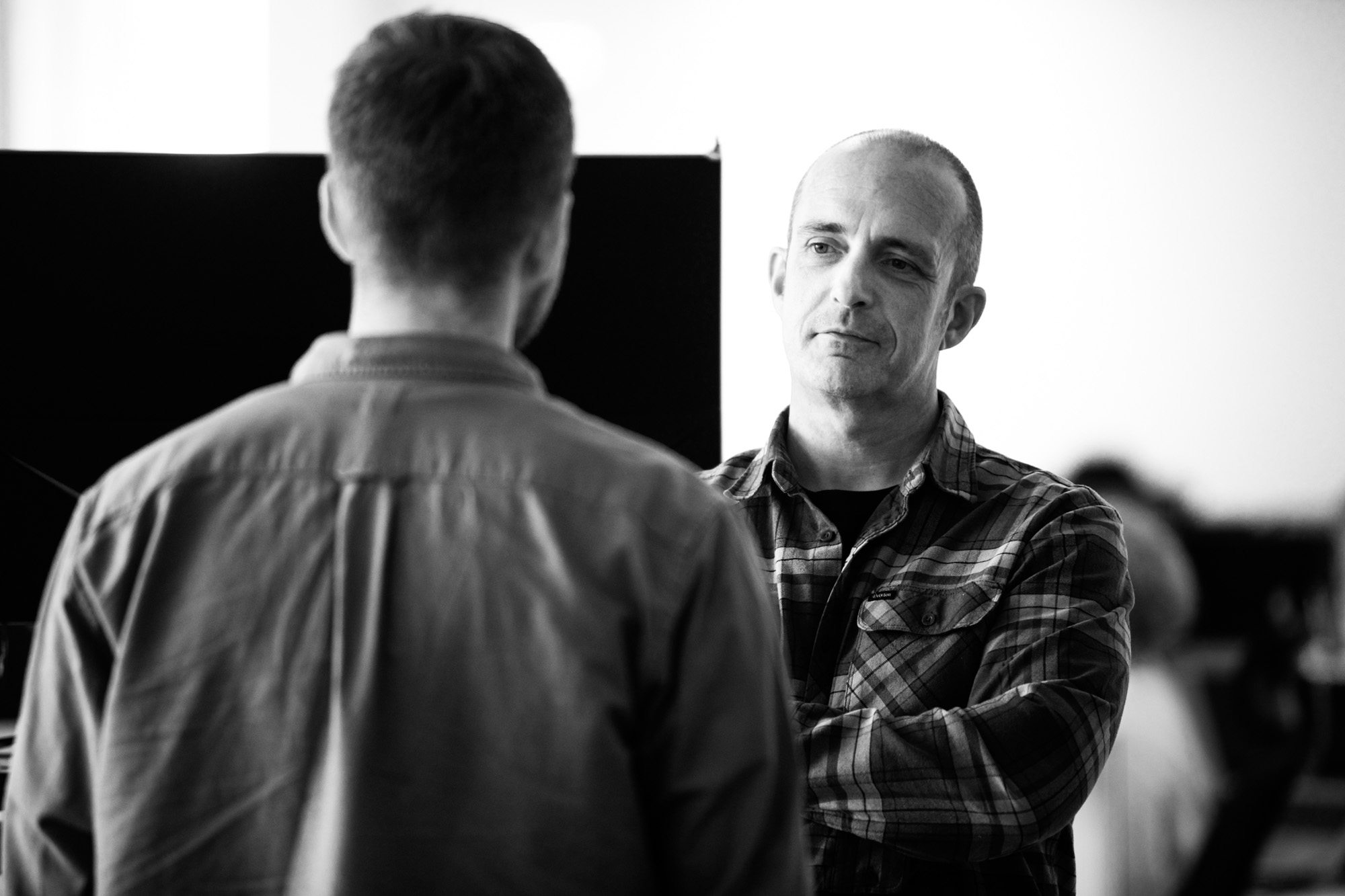 Two men in conversation, one facing away wearing light shirt, other facing forward in plaid shirt with arms crossed