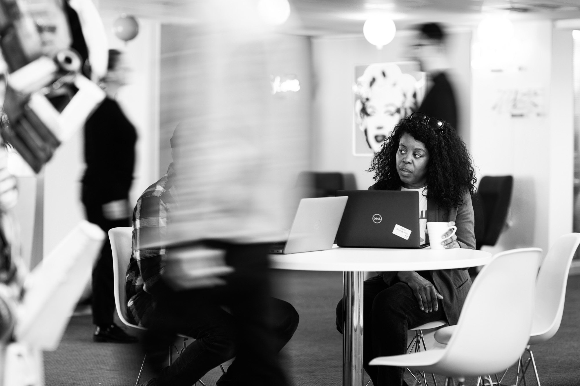 Woman with curly hair working on laptop at white table in modern office space with coffee cup