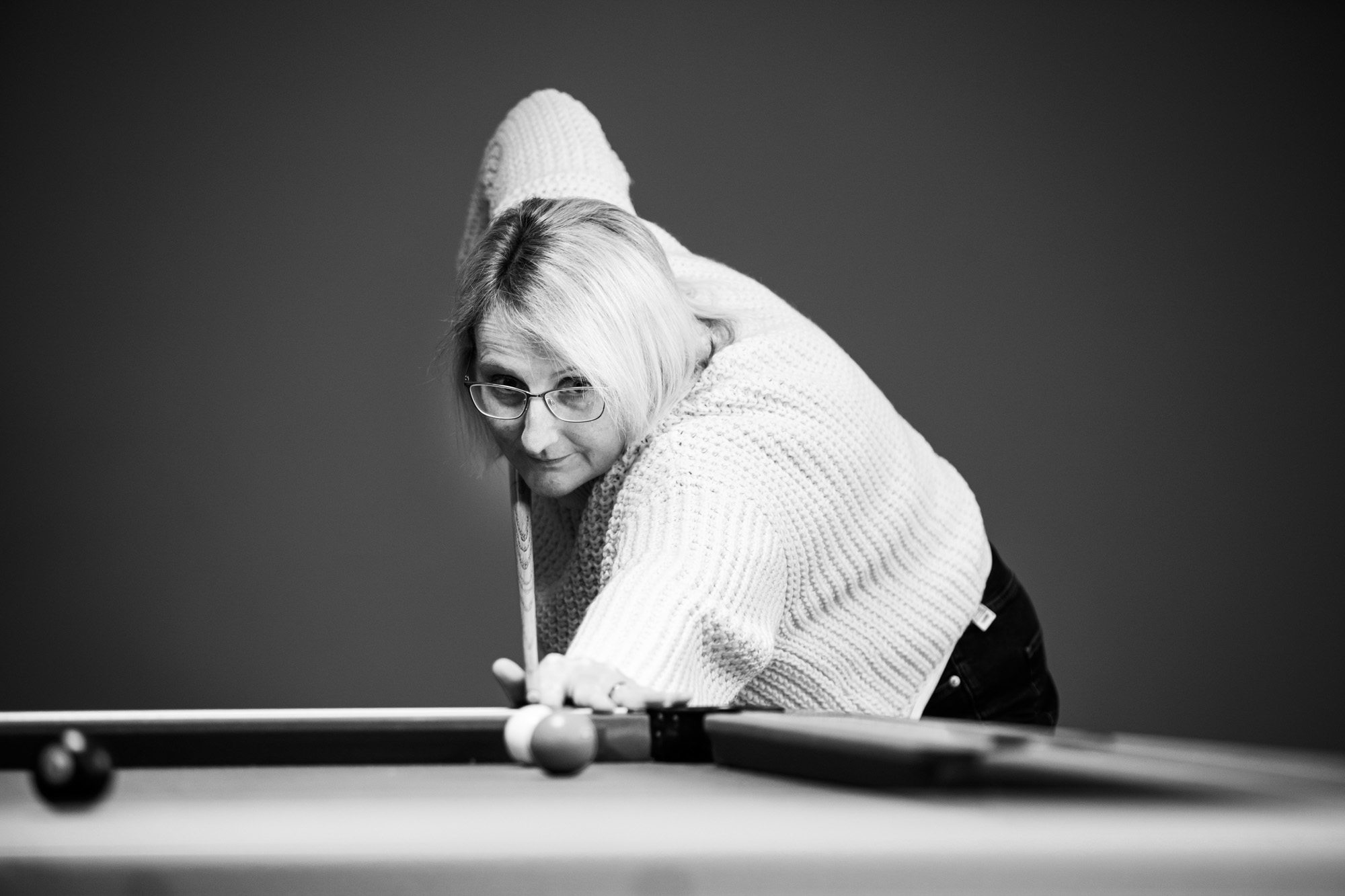Woman in knit sweater and glasses leaning over pool table with cue stick in black and white photo
