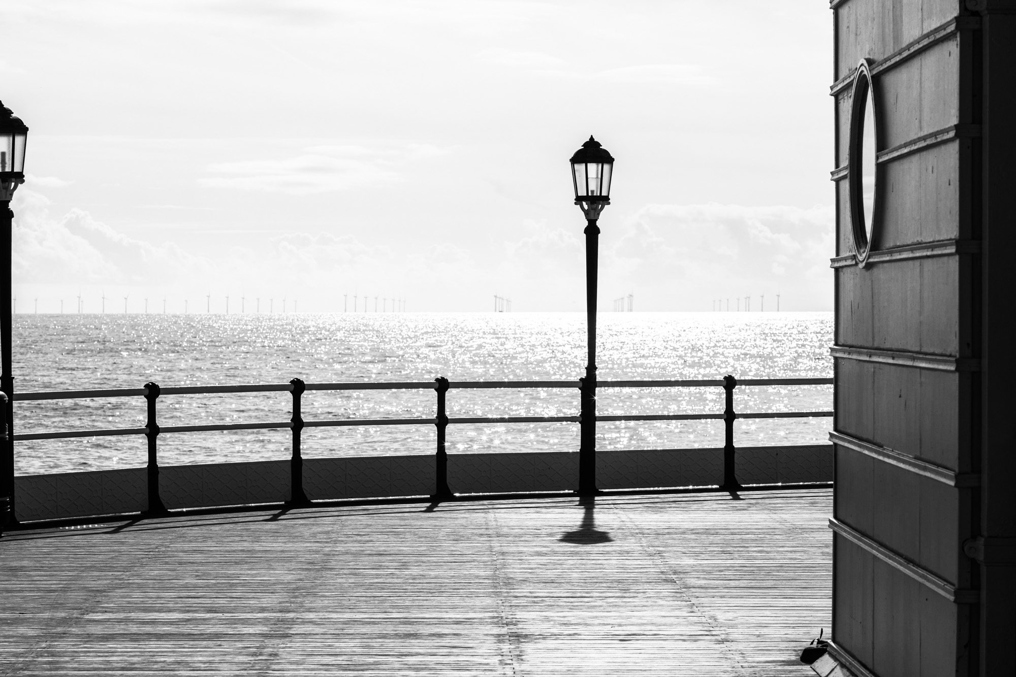Black and white view of wooden boardwalk with vintage lamp post and railing overlooking sparkling ocean water