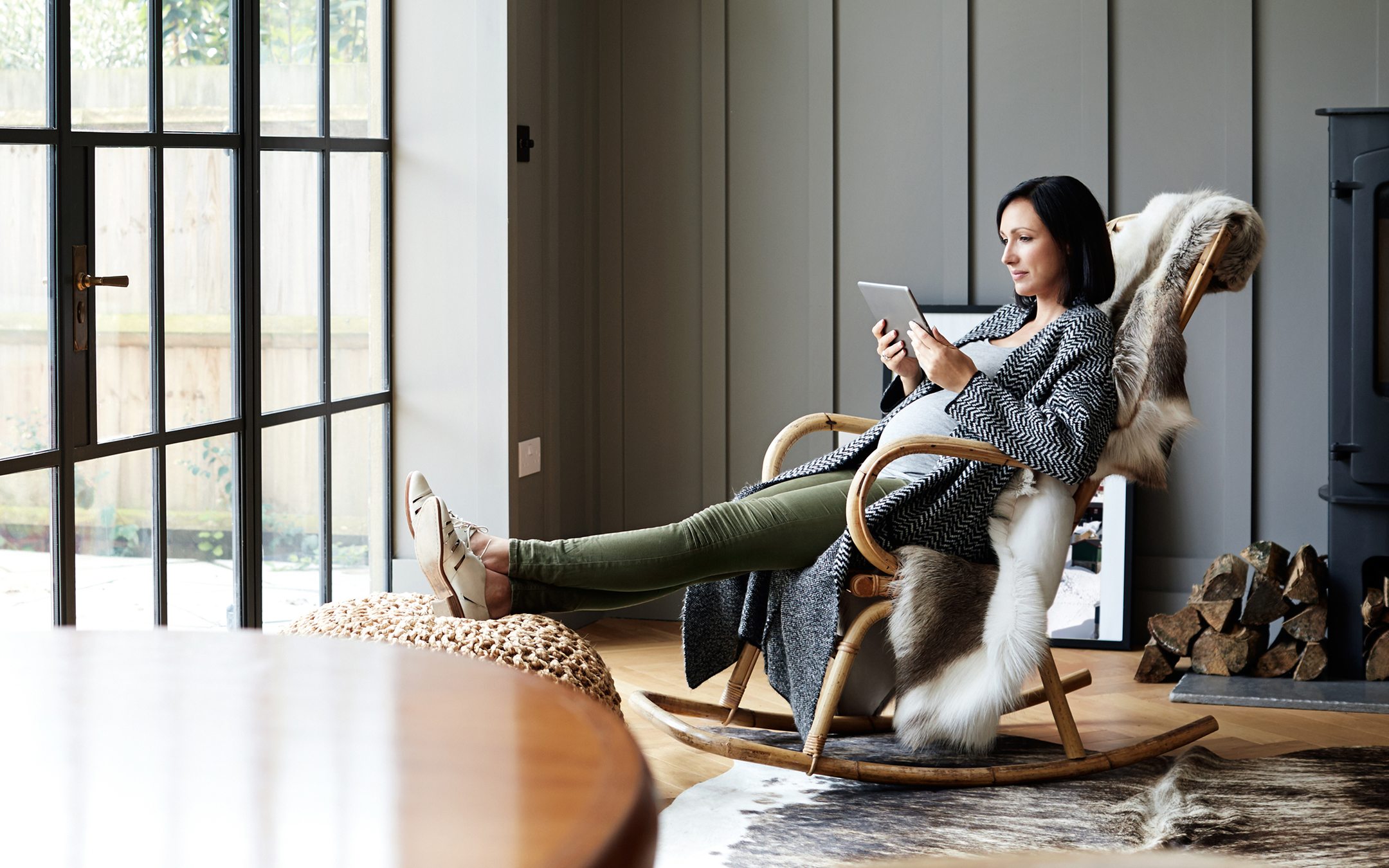 Woman relaxing in wooden rocking chair using tablet device in modern living room with large windows