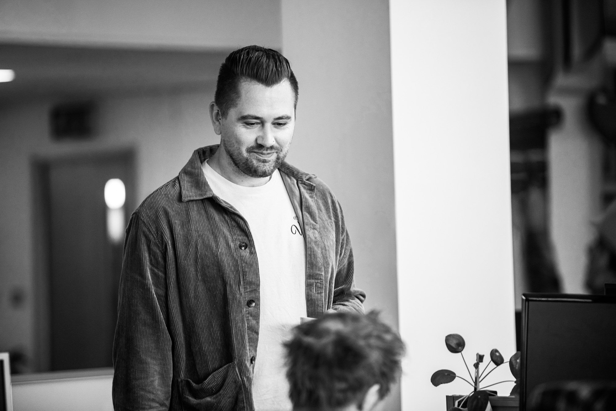 Man in corduroy jacket and white t-shirt smiling in modern office setting with blurred background