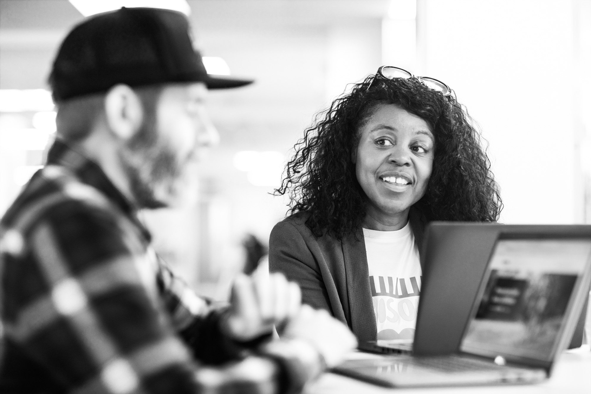 Woman with curly hair smiling at laptop computer in office workspace with male colleague in background
