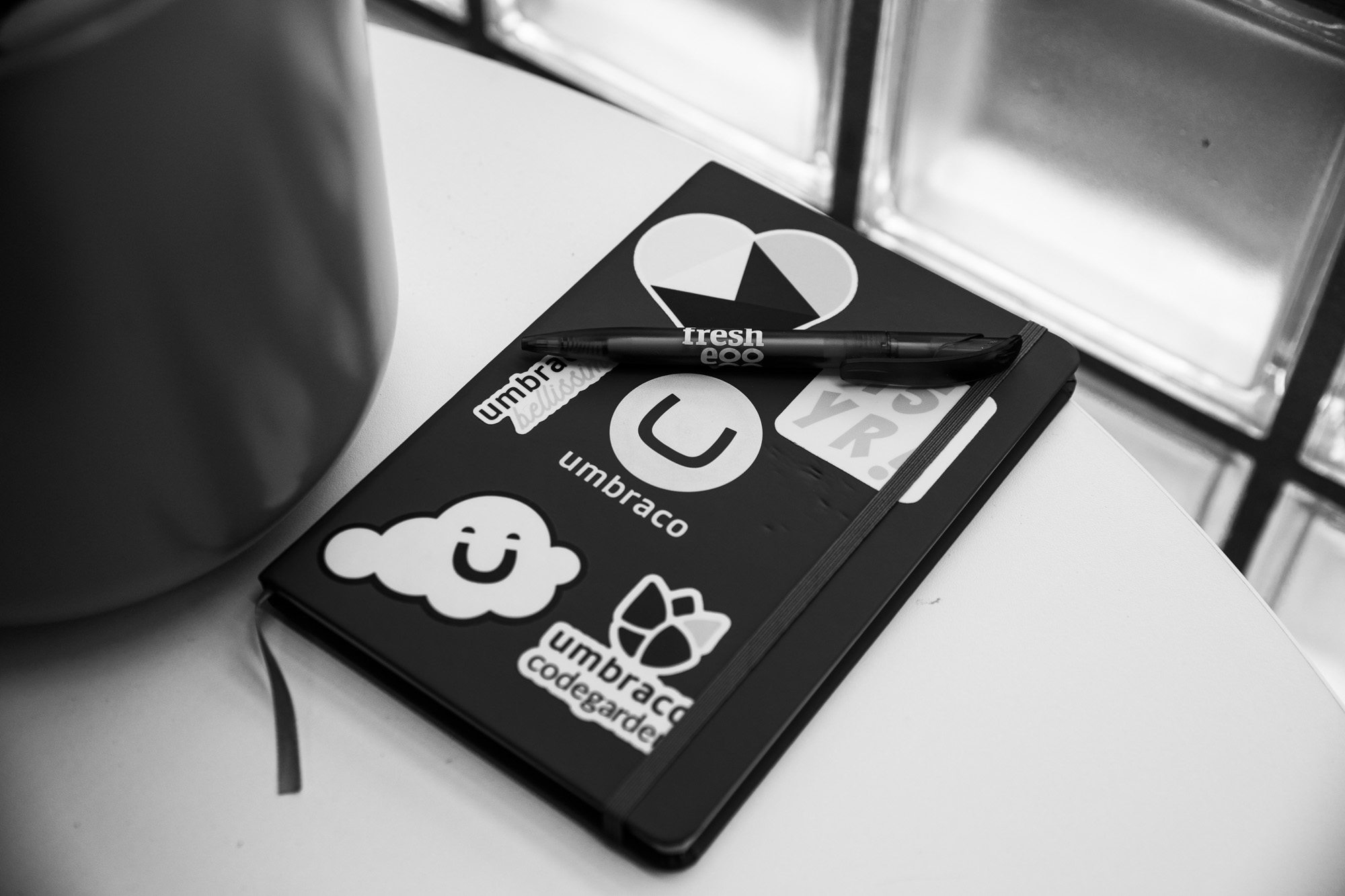 Black and white photo of laptop covered with tech company stickers including Umbreco, Fresh Eco logos on desk