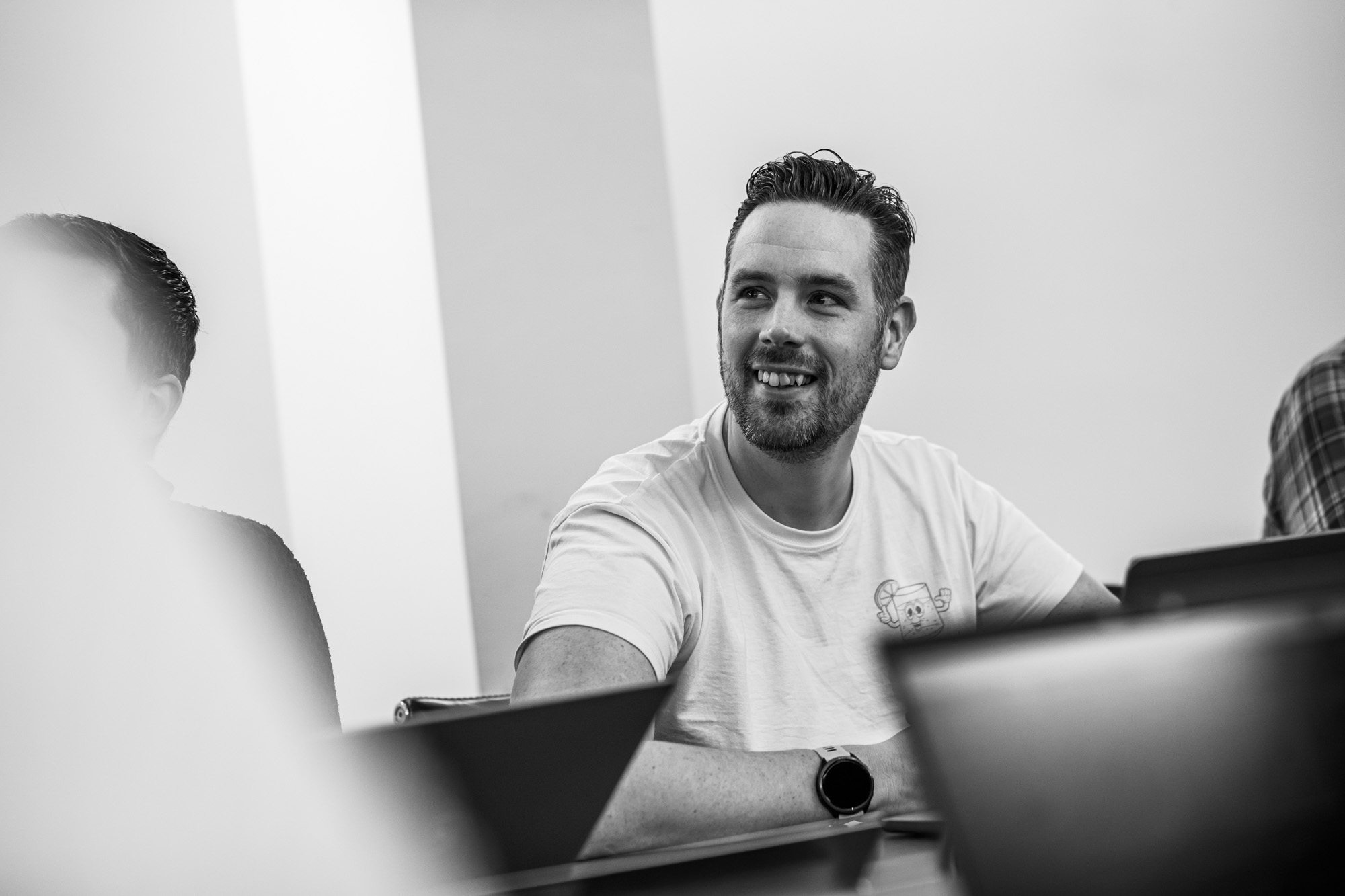 Smiling man with beard in white t-shirt sitting at desk with laptop in modern office setting, black and white photo