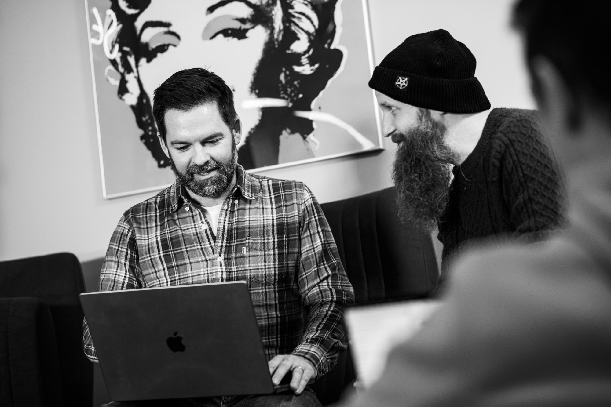 Two men collaborating at laptop in modern office with black and white portrait artwork on wall