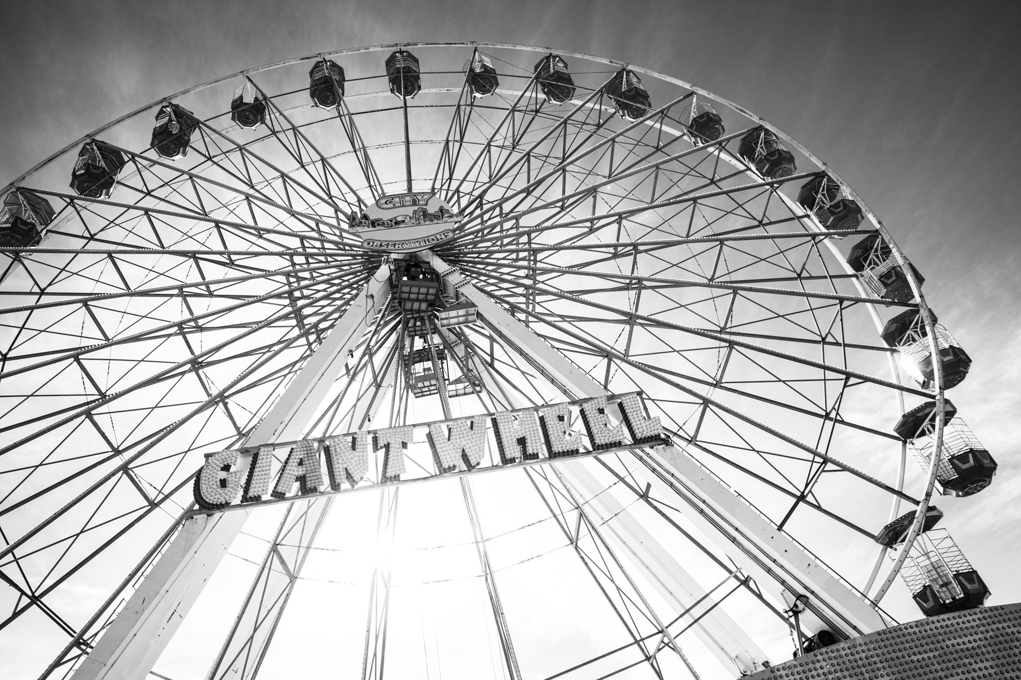 Black and white photograph of a large Ferris wheel shot from below against a cloudy sky