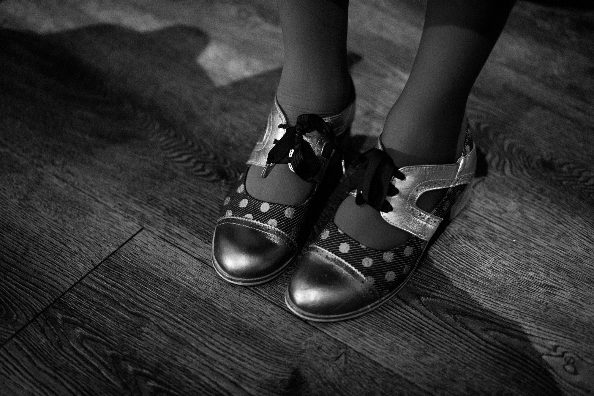 Black and white photo of child's feet wearing polka dot mary jane shoes on wooden floor