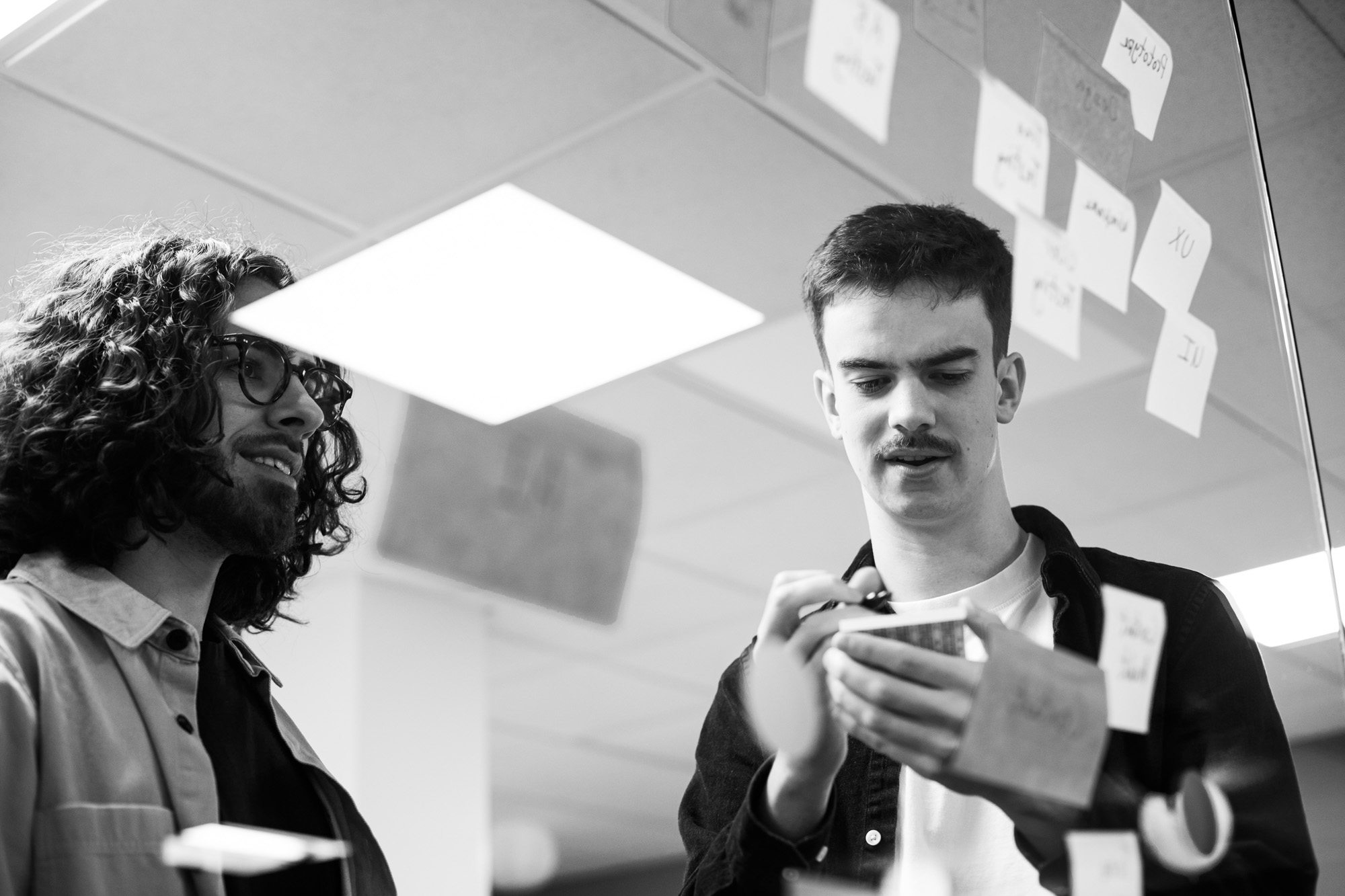 Two men collaborating in modern office, one with curly hair and glasses looking at colleague using smartphone