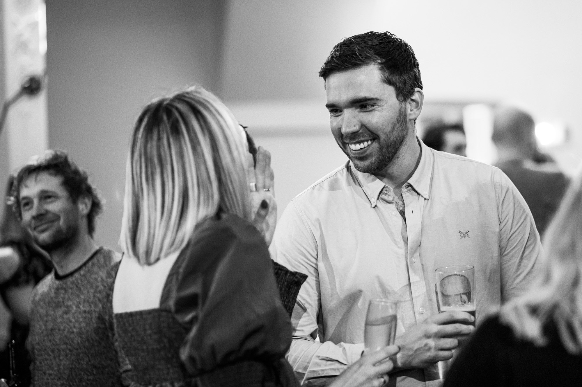 Smiling man in button-up shirt holding drink talking to blonde woman at social gathering, black and white photo