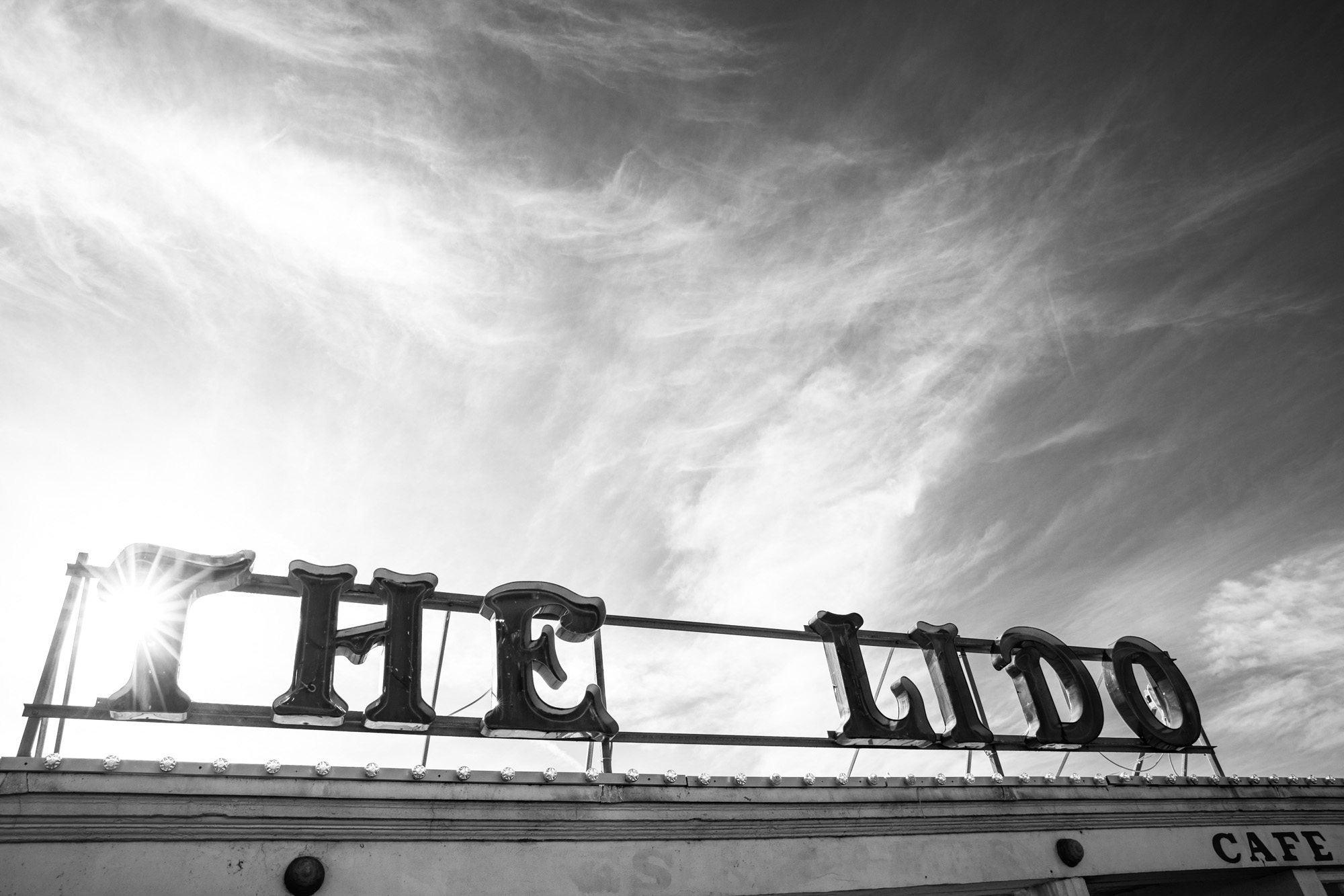 Black and white photo of vintage neon sign letters spelling "HUG LIDO" against dramatic cloudy sky with sun rays