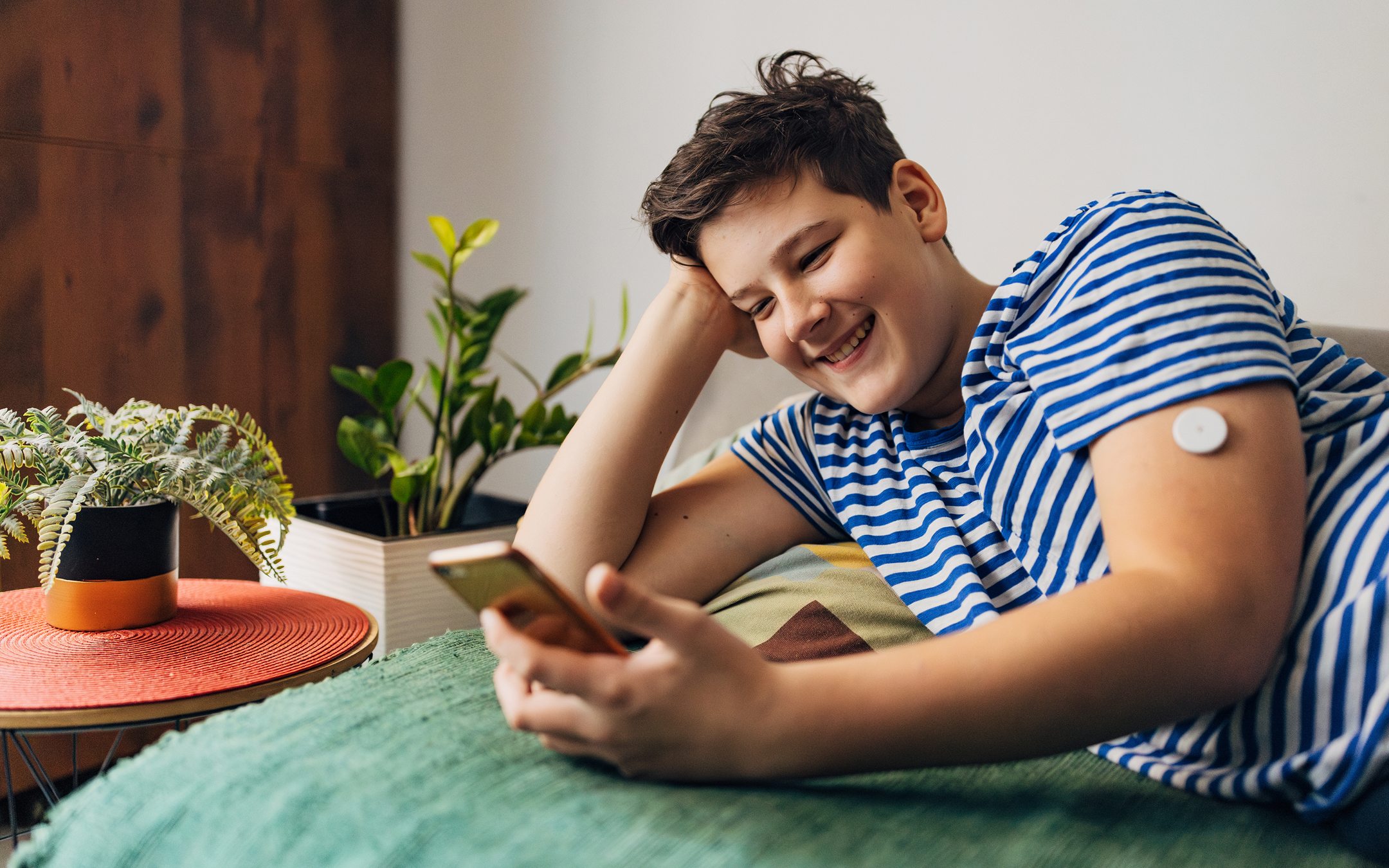 Young person with diabetes sensor on arm smiling while using smartphone, relaxing on green bedding with plants nearby